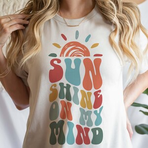 May include: A white t-shirt with a retro-style graphic design featuring a sun with rays and the text "SUNSHINE ON MY MIND" in colorful, wavy letters.