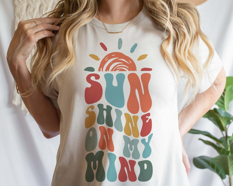 May include: A white t-shirt with a retro-style graphic design featuring a sun with rays and the text "SUNSHINE ON MY MIND" in colorful, wavy letters.