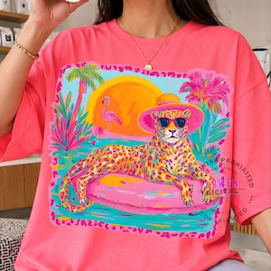 K&ouml;nnte beinhalten: Hellrosa T-Shirt mit einer farbenfrohen Illustration eines Leoparden mit Sonnenbrille und Hut, der auf einem rosa Schwimmreifen liegt. Die Grafik zeigt einen Sonnenuntergang, Palmen und einen Flamingo. Ein lebendiges, auff&auml;lliges Design.
