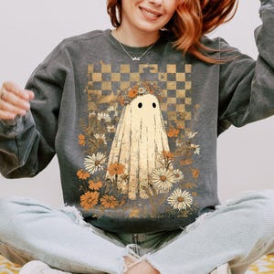 Op de afbeelding: Donkergrijze sweatshirt met een spookontwerp. De geest is cr&egrave;mekleurig met zwarte ogen, omringd door bloemen in tinten wit, oranje en bruin. Een goud-bruin geblokt patroon staat op de achtergrond. De sweatshirt is een casual, comfortabele stijl.