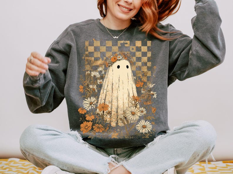 Op de afbeelding: Donkergrijze sweatshirt met een spookontwerp. De geest is cr&egrave;mekleurig met zwarte ogen, omringd door bloemen in tinten wit, oranje en bruin. Een goud-bruin geblokt patroon staat op de achtergrond. De sweatshirt is een casual, comfortabele stijl.