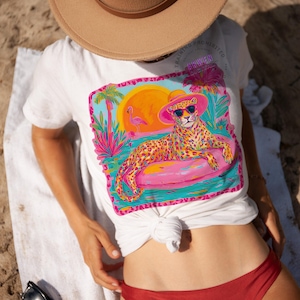 K&ouml;nnte beinhalten: Wei&szlig;es T-Shirt mit einem farbenfrohen Aufdruck eines Leoparden mit Sonnenbrille und rosa Hut, der auf einem rosa aufblasbaren Ring liegt. Das Design umfasst einen Sonnenuntergang, Palmen und einen Flamingo. Das T-Shirt ist an der Taille geknotet.