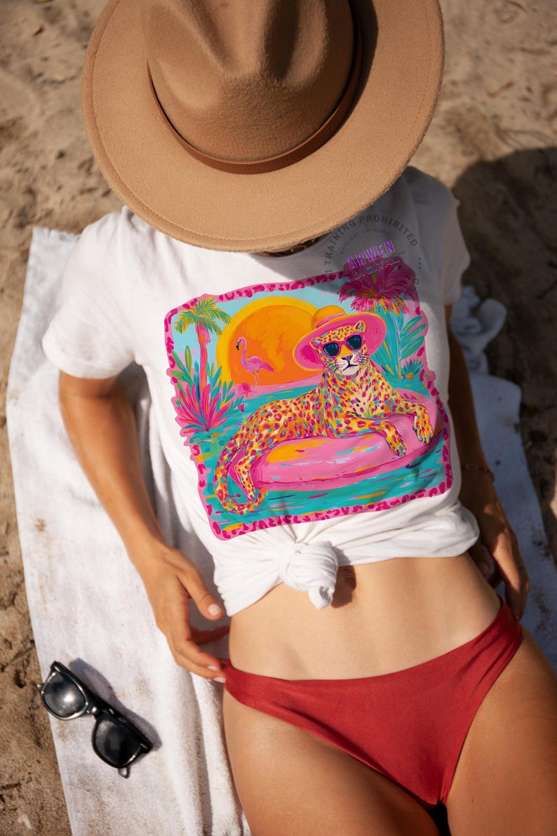 K&ouml;nnte beinhalten: Wei&szlig;es T-Shirt mit einem farbenfrohen Aufdruck eines Leoparden mit Sonnenbrille und rosa Hut, der auf einem rosa aufblasbaren Ring liegt. Das Design umfasst einen Sonnenuntergang, Palmen und einen Flamingo. Das T-Shirt ist an der Taille geknotet.