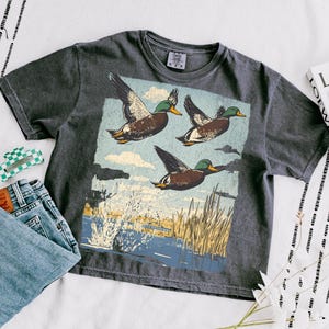 May include: Dark gray t-shirt featuring a graphic of three flying ducks over a marsh scene. The shirt is paired with blue jeans, a checkered hair clip, and a book. The shirt has a vintage, worn look.