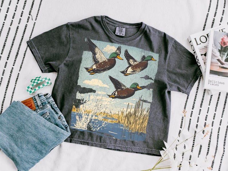 May include: Dark gray t-shirt featuring a graphic of three flying ducks over a marsh scene. The shirt is paired with blue jeans, a checkered hair clip, and a book. The shirt has a vintage, worn look.