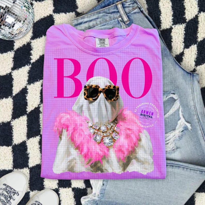 May include: A bright pink t-shirt featuring a large graphic of a ghost wearing leopard print sunglasses and a pink feather boa with a crystal statement necklace. The word BOO is printed in large pink letters above the ghost. The shirt has a tag.