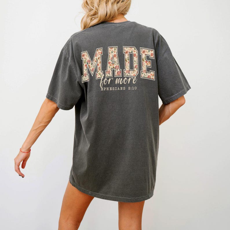 Op de afbeelding: Donkergrijs t-shirt met de tekst "MADE for more" in een bloemenpatroon op de rug. Daaronder staat de tekst "Ephesians 2:10" afgedrukt. Het shirt heeft een losse, comfortabele pasvorm.