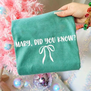 Może przedstawiać: Turkusowa koszulka z białym napisem &bdquo;MARY, DID YOU KNOW?&rdquo; i białą grafiką kokardy. Koszulka jest trzymana przed r&oacute;żową choinką z dekoracjami.