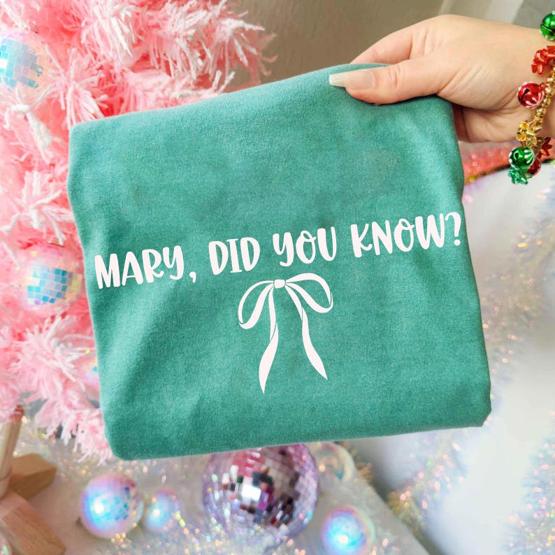 Może przedstawiać: Turkusowa koszulka z białym napisem &bdquo;MARY, DID YOU KNOW?&rdquo; i białą grafiką kokardy. Koszulka jest trzymana przed r&oacute;żową choinką z dekoracjami.