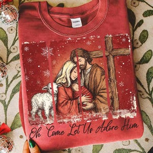 May include: A red sweatshirt featuring a religious illustration. The design includes a family with a baby, a lamb, and a cross. The text "Oh Come Let Us Adore Him" is printed at the bottom of the design.