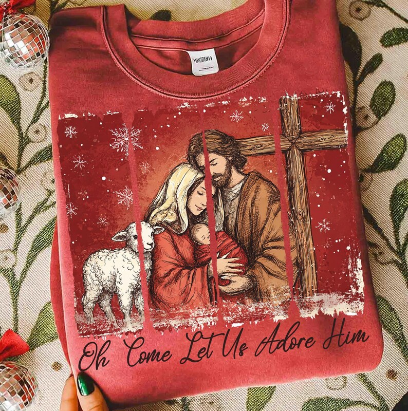 May include: A red sweatshirt featuring a religious illustration. The design includes a family with a baby, a lamb, and a cross. The text "Oh Come Let Us Adore Him" is printed at the bottom of the design.