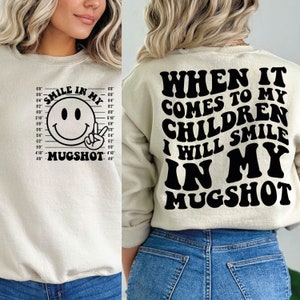 Può includere: Una felpa beige con una grafica nera sul davanti che dice "Smile in my Mugshot" con un viso sorridente all'interno di una cornice di foto segnaletica. La parte posteriore della felpa ha una grafica nera che dice "When it comes to my children I will smile in my mugshot".