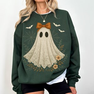 May include: Dark green sweatshirt featuring a ghost design with a bow, bats, and floral accents. The ghost is depicted with a lace-like appearance. The design is set against a dark green background, creating a festive Halloween theme.