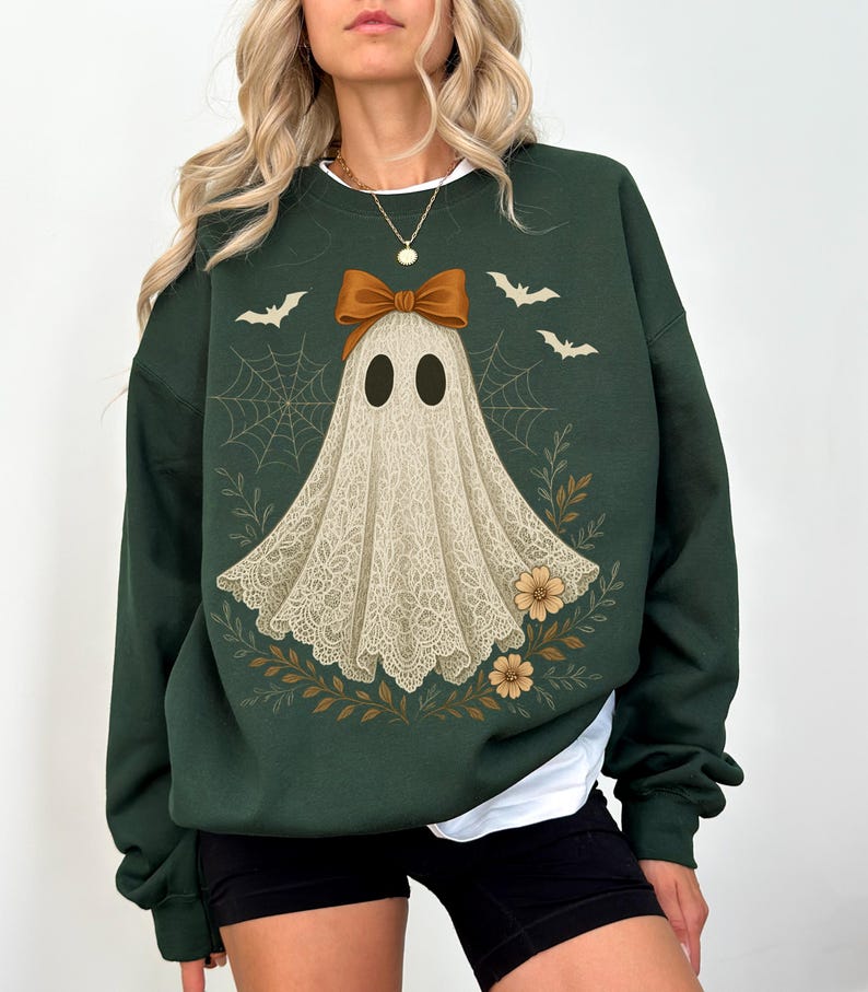 May include: Dark green sweatshirt featuring a ghost design with a bow, bats, and floral accents. The ghost is depicted with a lace-like appearance. The design is set against a dark green background, creating a festive Halloween theme.
