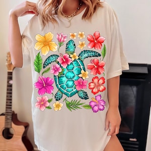 May include: White t-shirt featuring a colourful graphic of a sea turtle surrounded by tropical flowers. The turtle is teal blue and green with a detailed shell. The flowers are pink, red, yellow, and orange.