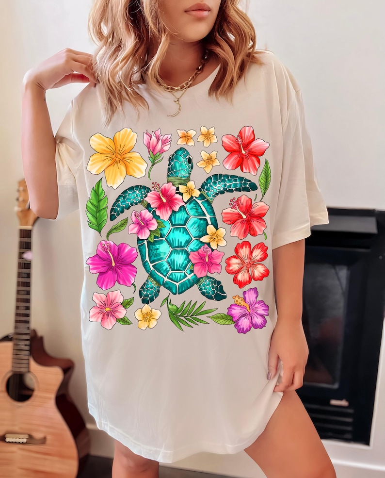 May include: White t-shirt featuring a colourful graphic of a sea turtle surrounded by tropical flowers. The turtle is teal blue and green with a detailed shell. The flowers are pink, red, yellow, and orange.