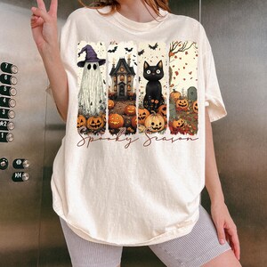 May include: Cream-colored t-shirt featuring a Halloween-themed graphic. The design includes a ghost, haunted house, black cat, pumpkins, and the words "Spooky Season". The shirt has a relaxed fit.