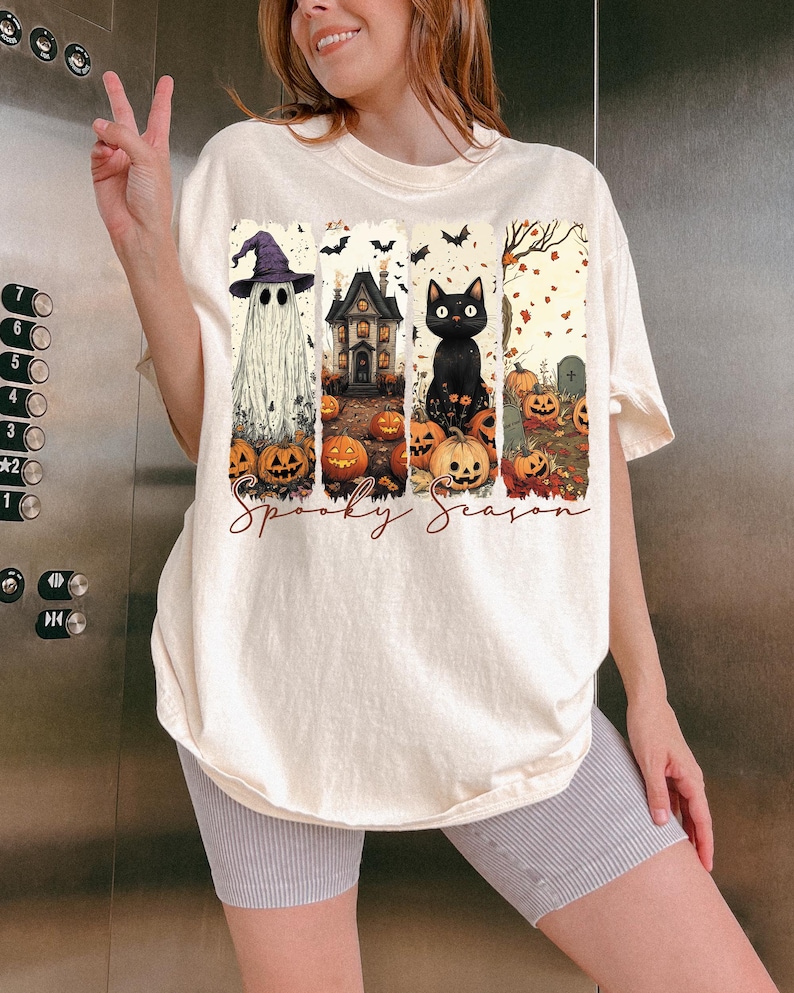 May include: Cream-colored t-shirt featuring a Halloween-themed graphic. The design includes a ghost, haunted house, black cat, pumpkins, and the words "Spooky Season". The shirt has a relaxed fit.