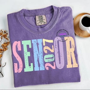 Puede incluir: Una camiseta morada de manga corta con un estampado gr&aacute;fico desgastado que dice SENIOR 2027 en letras de bloque de colores pastel rosa, verde, azul, amarillo y lavanda. La camiseta tiene una etiqueta de la marca Comfort Colors en el cuello.