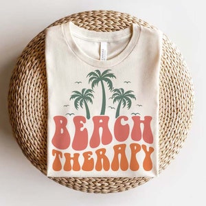 May include: A beige t-shirt with a retro-style graphic of palm trees and the text "Beach Therapy" in orange and pink.