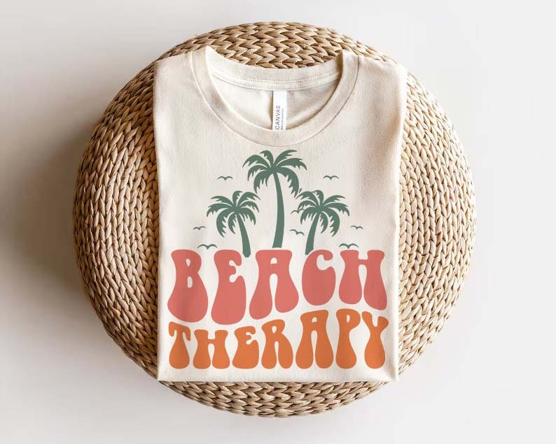 May include: A beige t-shirt with a retro-style graphic of palm trees and the text "Beach Therapy" in orange and pink.