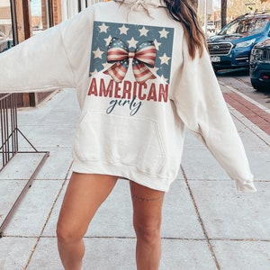 May include: A cream-colored hoodie with a vintage-style American flag bow and the text "AMERICAN girly".