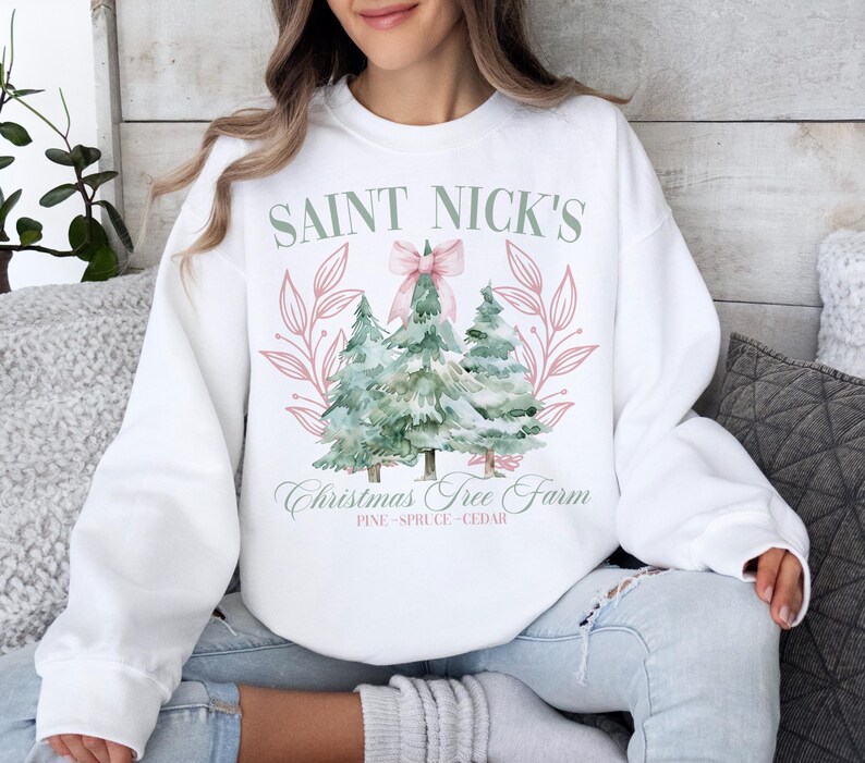 May include: White sweatshirt with a watercolor design featuring three Christmas trees, a pink bow, and the text "Saint Nick's Christmas Tree Farm." The design includes the words "Pine, Spruce, Cedar."