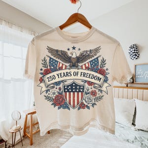 May include: Cream-colored t-shirt with a patriotic design. The graphic features an eagle, American flags, and a shield with stars and stripes, surrounded by roses and the text "250 YEARS OF FREEDOM".