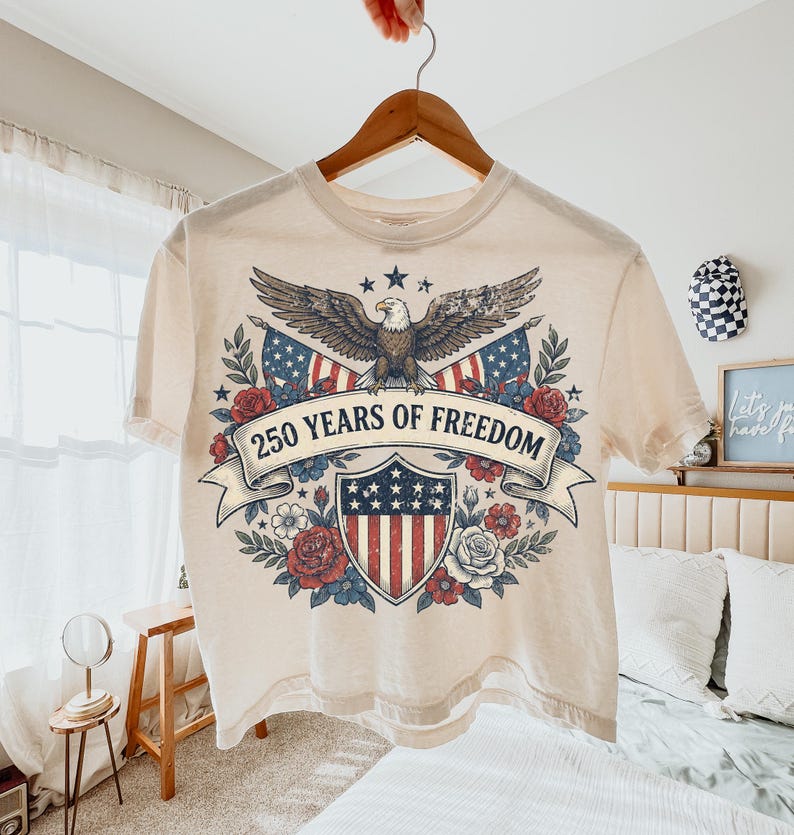 May include: Cream-colored t-shirt with a patriotic design. The graphic features an eagle, American flags, and a shield with stars and stripes, surrounded by roses and the text "250 YEARS OF FREEDOM".