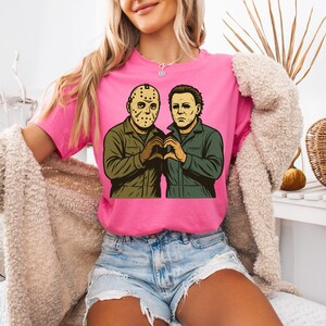 May include: A pink t-shirt featuring a graphic of two horror movie characters, Jason Voorhees and Michael Myers, forming a heart shape with their hands. The characters are depicted in a cartoon style with green and brown clothing.