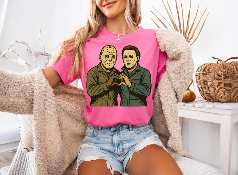May include: A pink t-shirt featuring a graphic of two horror movie characters, Jason Voorhees and Michael Myers, forming a heart shape with their hands. The characters are depicted in a cartoon style with green and brown clothing.