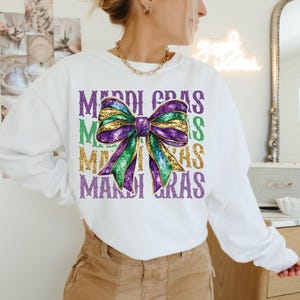 Puede incluir: Sudadera blanca con un lazo de purpurina morado, verde y dorado y el texto "Mardi Gras" repetido cuatro veces.