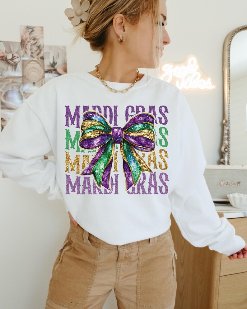 Puede incluir: Sudadera blanca con un lazo de purpurina morado, verde y dorado y el texto "Mardi Gras" repetido cuatro veces.