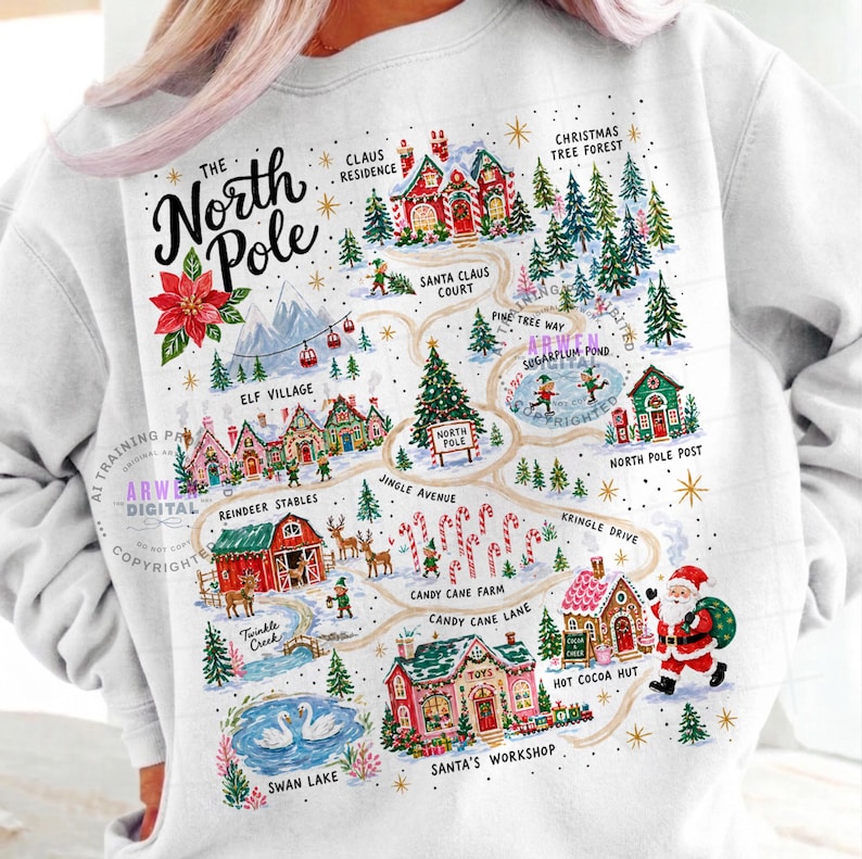 May include: A white sweatshirt featuring a colorful graphic map of the North Pole. The design includes illustrations of Santa's workshop, reindeer stables, and candy cane lanes, with text labels for each location and a small Santa Claus figure.