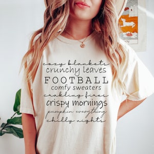 May include: Cream-colored t-shirt with black script text listing fall-themed phrases: cozy blankets, crunchy leaves, football, comfy sweaters, crackling fires, crispy mornings, pumpkin everything, and chilly nights.