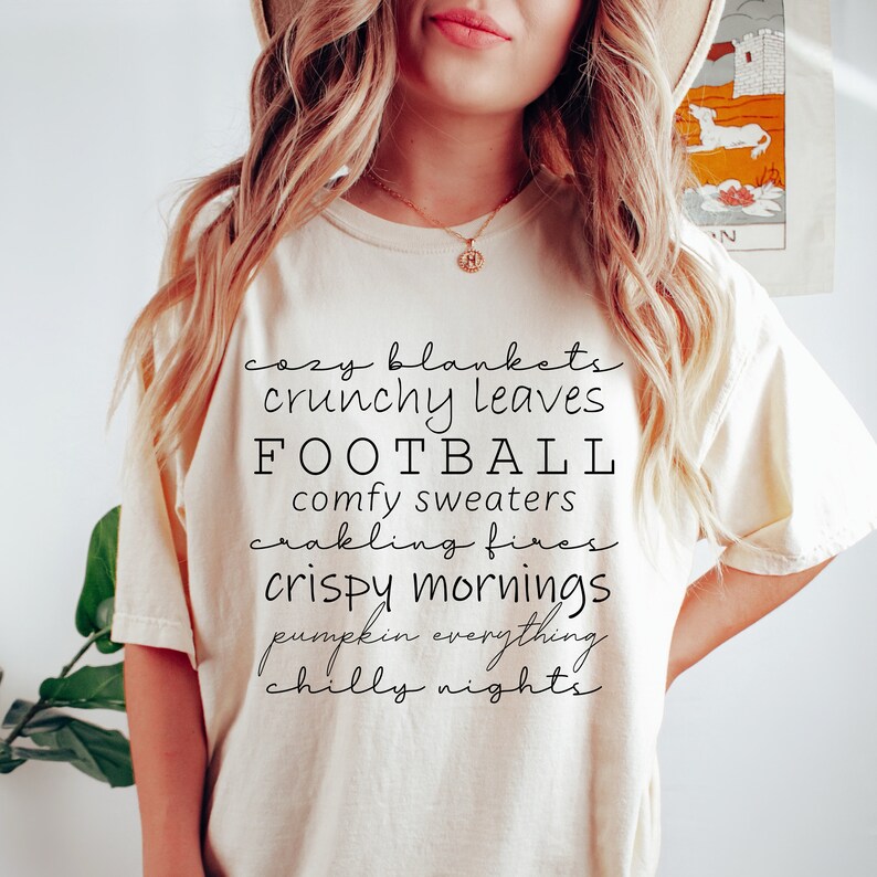 May include: Cream-colored t-shirt with black script text listing fall-themed phrases: cozy blankets, crunchy leaves, football, comfy sweaters, crackling fires, crispy mornings, pumpkin everything, and chilly nights.