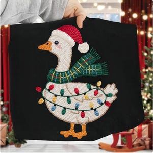 May include: Embroidered black pillow featuring a festive goose wearing a Santa hat and a green scarf. The goose is adorned with a string of colorful Christmas lights, creating a whimsical holiday design.
