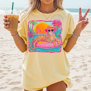 K&ouml;nnte beinhalten: Ein gelbes T-Shirt mit einem farbenfrohen Grafikdesign. Das Design zeigt einen Leoparden mit Sonnenbrille und rosa Hut, der auf einem rosa aufblasbaren Ring liegt. Das Design umfasst einen Sonnenuntergang, Palmen und einen Flamingo. Der Text "TRAINING PROHIBITED" ist ebenfalls sichtbar.