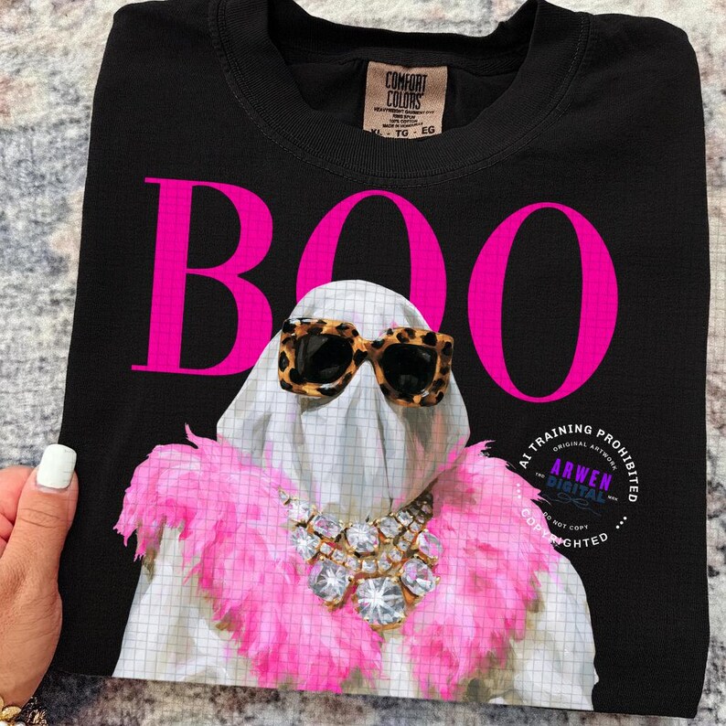 May include: A black t-shirt featuring the word BOO in large hot pink letters. The graphic shows a white ghost wearing leopard print sunglasses, a pink feather boa, and a large crystal statement necklace. The shirt has a Comfort Colors brand label.