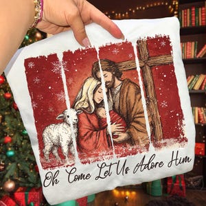 May include: White pillow with a religious illustration featuring a family, a lamb, and a cross, set against a red background with snowflakes. The text "Oh Come Let Us Adore Him" is at the bottom.