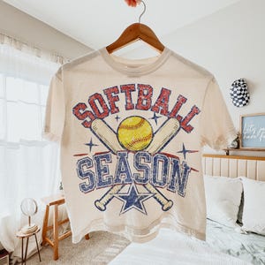 May include: Cream-colored t-shirt with the words "Softball Season" in red and blue distressed lettering. The design includes a yellow softball, crossed baseball bats, and a blue star. The shirt is hanging on a wooden hanger.