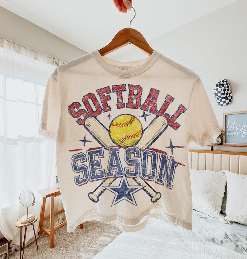 May include: Cream-colored t-shirt with the words "Softball Season" in red and blue distressed lettering. The design includes a yellow softball, crossed baseball bats, and a blue star. The shirt is hanging on a wooden hanger.