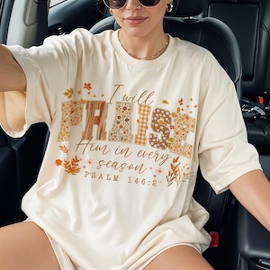 May include: Cream-coloured t-shirt featuring the phrase "I will PRAISE Him in every season" in various patterned fonts. The text includes "PSALM 146:2" with autumn-themed leaf and floral accents. The shirt is a relaxed fit.