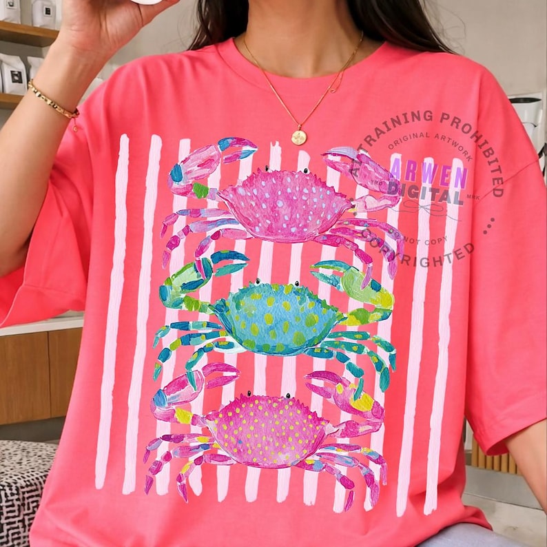Puede incluir: Una camiseta de color coral con un dise&ntilde;o caprichoso de tres cangrejos coloridos. Los cangrejos son en tonos de rosa, azul y verde, con un patr&oacute;n de puntos. Rayas blancas verticales recorren la camiseta. El texto "ARWEN DIGITAL" es visible.