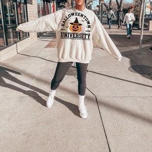 May include: A cream-coloured sweatshirt featuring the words "Halloweentown University" and a jack-o'-lantern graphic. The sweatshirt has long sleeves and a crew neck. The text "Where being normal is vastly overrated" is printed below the university name.