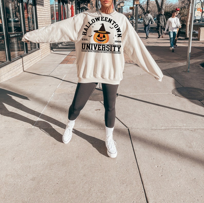 May include: A cream-coloured sweatshirt featuring the words "Halloweentown University" and a jack-o'-lantern graphic. The sweatshirt has long sleeves and a crew neck. The text "Where being normal is vastly overrated" is printed below the university name.