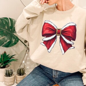 Puede incluir: Sudadera beige de cuello redondo con un gran dise&ntilde;o de lazo rojo con ribete blanco y hebilla dorada. El lazo es un gr&aacute;fico festivo, perfecto para la temporada navide&ntilde;a. La sudadera es de color neutro, adecuada para varios conjuntos.