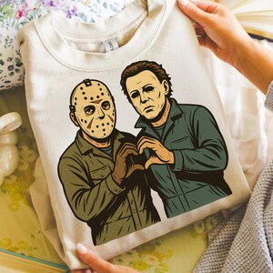 May include: A cream-colored sweatshirt featuring a graphic of two horror movie characters, Jason Voorhees and Michael Myers, forming a heart shape with their hands. The artwork is in a vintage comic book style, with the characters in their signature outfits.