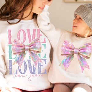 K&ouml;nnte beinhalten: Zwei wei&szlig;e Sweatshirts mit einem rosafarbenen und schillernden Schleifendesign. Das gr&ouml;&szlig;ere Sweatshirt tr&auml;gt den Text "LOVE LIKE LOVE like jesus" in pinkfarbenem, gr&uuml;nem und violettem Glitzer.
