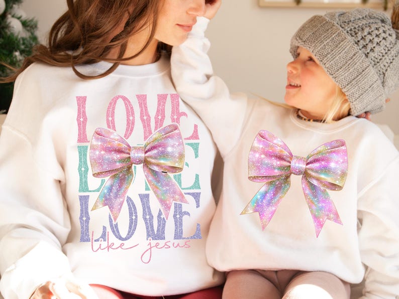 K&ouml;nnte beinhalten: Zwei wei&szlig;e Sweatshirts mit einem rosafarbenen und schillernden Schleifendesign. Das gr&ouml;&szlig;ere Sweatshirt tr&auml;gt den Text "LOVE LIKE LOVE like jesus" in pinkfarbenem, gr&uuml;nem und violettem Glitzer.